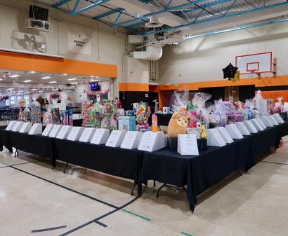 Indoor event with decorated tables displaying raffle prizes, toys, and gift baskets in a gymnasium setting.