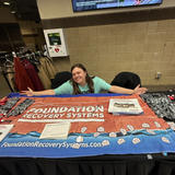 Person smiling at a booth displaying "Foundation Recovery Systems" with brochures and promotional items.
