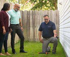 An inspector kneels on grass showing equipment to two people by a house and wooden fence.