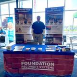 A man stands behind a Foundation Recovery Systems booth with informational displays and brochures on the table.