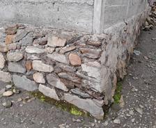 A stone and concrete building foundation with moss growing at the base and loose rocks nearby.