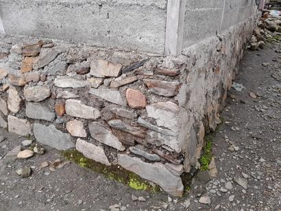A stone and concrete building foundation with moss growing at the base and loose rocks nearby.