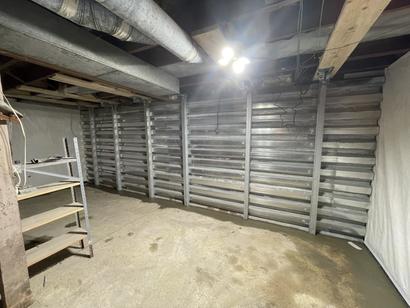 Unfinished basement with metal support beams on the wall, exposed ceiling pipes, and a metal shelving unit.