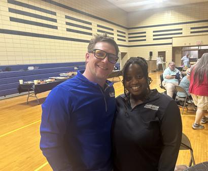 Two people smiling and posing together in a gymnasium with tables and people in the background.