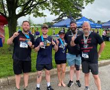 Five smiling runners pose with medals and race bibs, holding up one finger, at an outdoor event.