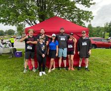 Seven people in race attire stand smiling in front of a red tent on a grassy area at an outdoor event.