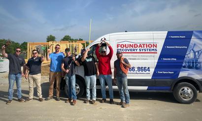 Seven construction workers pose with tools in front of a Foundation Recovery Systems van at a building site.