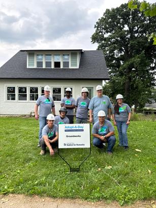 Eight people in hard hats pose outside a house with a Habitat for Humanity Adopt-A-Day sign in front.