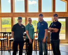 Four people stand smiling indoors in front of large windows with a scenic outdoor view.