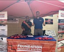 Two men stand at a Foundation Recovery Systems booth under a red canopy, smiling and giving thumbs up.