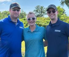 Three people in sunglasses and caps smiling outdoors, standing together in front of trees and greenery.