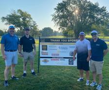 Four men stand on grass near a sponsor sign at a golf event on a sunny day with trees in the background.
