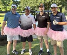 Four men outdoors, three wearing pink tutus and golf attire, smiling for a group photo by a golf cart.