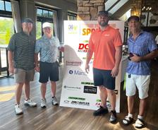 Four men standing indoors next to a sponsor sign at a golf event, smiling at the camera.