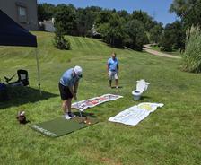 Two men play mini-golf in a yard, aiming at a toilet; towels and a mat are on the grass.