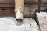 Beige downspout draining onto snowy ground beside a stone wall and steps.
