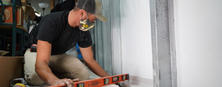 A person wearing a cap and mask uses a level tool to align a metal frame indoors.