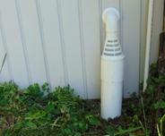 White vent pipe with a conical top next to a building wall, surrounded by grass and leaves.