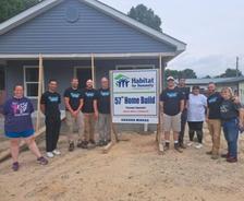 A group poses by a Habitat for Humanity sign in front of a newly-built house on a dirt lot.