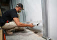 A construction worker measuring a basement wall with a level tool, wearing a mask and cap.