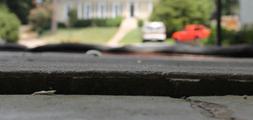 Close-up of a cracked pavement with a blurred view of a house, white car, and red truck in the background.