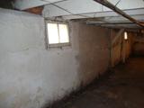 A dimly lit, empty basement with exposed beams and small windows casting light on cracked, white-painted walls.
