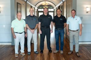 Five men standing in a hallway, smiling and dressed in casual business attire.