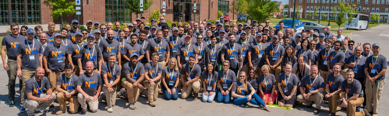 A large group of people in matching shirts pose together outside a building on a sunny day.