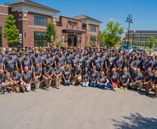 A large group of people in matching shirts pose together outside a building on a sunny day.