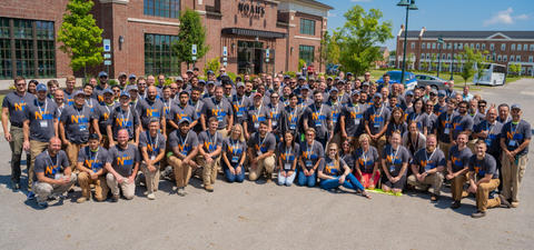 A large group of people in matching shirts pose together outside a building on a sunny day.