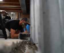 A person wearing a cap and mask crouches down indoors, carefully working on a metal beam as part of the foundation waterproofing process.