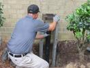 A man wearing gloves installs a foundation pier beside a brick wall, surrounded by small bushes.
