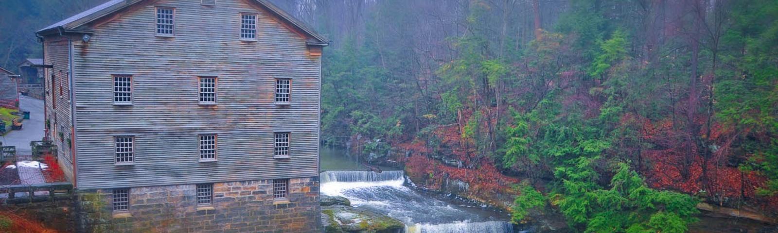 Historic wooden mill by a waterfall in a misty forest landscape.