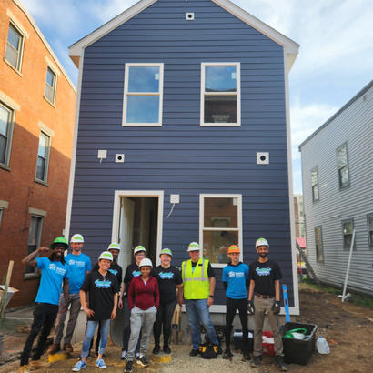A group of people wearing hard hats stands smiling in front of a newly built blue house.
