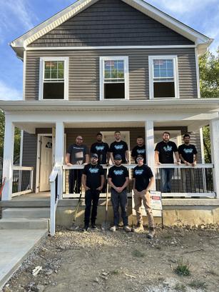 A group of nine people in matching shirts stands in front of a gray house with a porch.