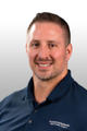 Man with short hair, beard, and blue polo shirt smiling against a light gray background.