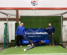 Three people pose in front of a "Basement Authority" banner on artificial grass, with one person lying down playfully.