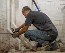 A plumber kneels to fix PVC pipes amidst tools, in a basement with white brick walls, ensuring excellent basement waterproofing.