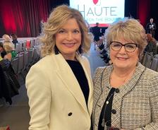 Two women smiling at a fashion event with a "HAUTE" sign in the background.