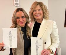 Two women smiling and holding fashion sketches, one in black and the other in white clothing, indoors.