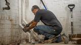 A plumber wearing gloves works on white pipes in a basement with brick walls and a shovel beside him.