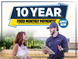 A man with a clipboard talks to a woman near a fence, with a banner reading "10 Year Fixed Monthly Payments!