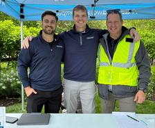 Three smiling men stand together under a blue canopy, one wearing a yellow safety vest, at an outdoor event.