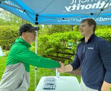 Two men shake hands under a blue tent at an outdoor event, standing by a table with packaged items.